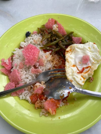 A close-up, overhead shot shows a plate of food on a white, textured surface. The plate is a vibrant lime green and contains a serving of white rice, topped with a fried egg and various savory dishes. Scattered throughout the rice are pieces of bright pink crackers, adding a pop of color. A fork and spoon rest on the plate, ready for consumption. The food appears to be a popular dish in Keuniree, Indonesia, known for its flavorful and aromatic cuisine. The lighting is bright, suggesting an outdoor setting or a well-lit indoor space.