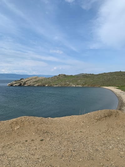 The image shows a natural landscape in the city of Sakhiurta, Russia. The foreground is dominated by a sandy embankment, sparsely dotted with small green plants and pebbles. Beyond this embankment, a calm, dark blue body of water stretches into the distance, meeting a curved, sandy shoreline. The shoreline is backed by a gently sloping, green, rocky hill. On the far left of the image, a rocky peninsula juts out into the water. The sky above is a pale blue, with wispy white clouds scattered across it. The overall scene depicts a serene, natural environment with a focus on the interplay between land, water, and sky.