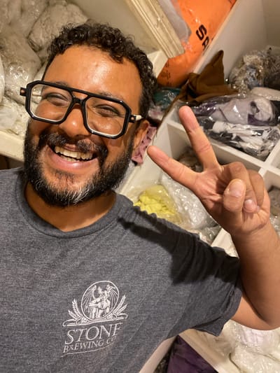 A man with dark curly hair and a beard, wearing black-rimmed glasses and a grey t-shirt with a "Stone Brewing Co." logo, smiles at the camera. He is making a peace sign with his right hand. The man appears to be in a storage area, with shelves filled with various items, including bags of fabric or material, and folded clothing. The background suggests an indoor environment, possibly a warehouse or a large closet, with shelves and possibly a window or light source visible. The man's t-shirt features a circular logo with an illustration of a creature and wheat stalks. The lighting is bright, and the image is a close-up shot focused on the man. No specific location in the city of Legenda, Egypt, can be determined from the visual information provided.
