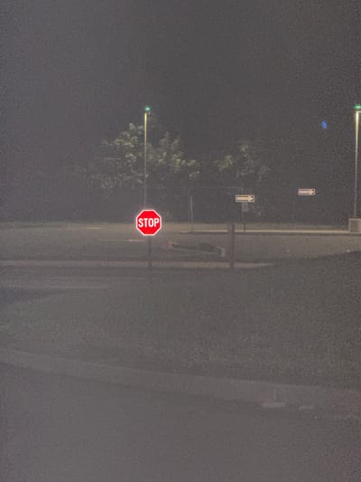The image depicts a nighttime scene at an intersection in New Britain, Connecticut, United States of America. A red, octagonal stop sign with white lettering reading "STOP" is prominently displayed in the foreground, illuminated by an unseen light source. Beyond the stop sign, two "ONE WAY" signs with white arrows on yellow backgrounds are visible, indicating traffic flow directions. Streetlights with green tops are present in the background, casting a dim glow on the surroundings. The environment appears to be an unlit roadway with grassy areas and trees, suggesting a suburban or possibly a park-like setting within the city. The overall atmosphere is hazy and dimly lit, consistent with a foggy or misty night. There are no people visible in the image.