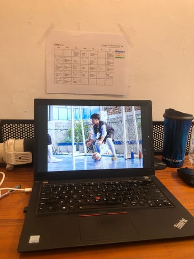 An overhead shot captures a workspace in Cibinong, Indonesia, featuring a black Lenovo ThinkPad laptop, a calendar, and various desk items on a wooden surface. The laptop is open, displaying a video of a young male figure on a blue futsal court, actively engaged in a game or practice with a soccer ball. He is wearing athletic gear including knee pads and white sneakers. The background of the video shows a net and hints of greenery, suggesting an outdoor or semi-outdoor court setting under daylight conditions.
To the left of the laptop, a digital calendar, labeled "Angga content planner," is taped to a textured, off-white wall. The calendar prominently displays "June 2026" and shows the days of the week from Sunday to Saturday, with dates ranging from May 31st to June 30th. Small, unreadable notes are present on several dates.
Behind the laptop, a blue reusable water bottle with a black lid stands upright, featuring the text "BLUE BIRD GROUP." A black computer mouse is partially visible. Beneath the laptop, a white device, possibly an adapter or power bank, is positioned.
Visible text includes "ThinkPad," "T480s," and an "Intel CORE i5" sticker on the laptop. The laptop's keyboard shows various letters and symbols. The calendar text includes "Angga content planner" and "June 2026." The water bottle clearly displays "BLUE BIRD GROUP." The overall scene suggests a personal or home office environment, with the laptop screen providing a dynamic focal point.