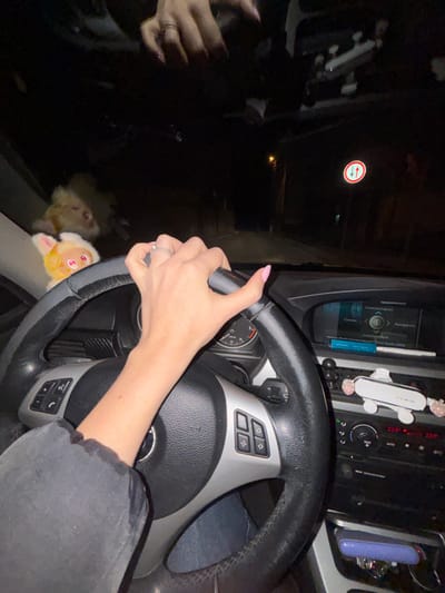 The image depicts the interior of a vehicle at night, specifically in Kisyovtsi, Bulgaria, from the driver's perspective. A person's right arm, clad in a grey long-sleeved garment, is visible, with the hand gripping the black leather steering wheel. The hand features pink manicured nails and a silver-colored ring on the ring finger. Reflections of another hand with similar nails are visible in the windshield above. The steering wheel, a BMW model, shows wear and has multifunction buttons on its spokes.
Through the windshield, a dark, poorly lit road extends forward. A street light illuminates a small section of the road in the distance on the right. A circular red road sign with two vertical arrows (one up, one down) is visible on the right side of the road. On the dashboard, two plush toys are visible: a larger, light-colored fluffy animal and a smaller orange-and-yellow creature with ears.
The vehicle's dashboard features a screen displaying "Communication," "Entertainment," and "Navigation." Below the screen are climate controls showing "23.5°C" on both sides. A white, rectangular phone holder with pink accents is mounted on the dashboard to the right of the steering column. A blue object is lodged in an air vent above the center console, and a purple-cased object is visible in a storage compartment below the climate controls. The driver is also wearing blue jeans.