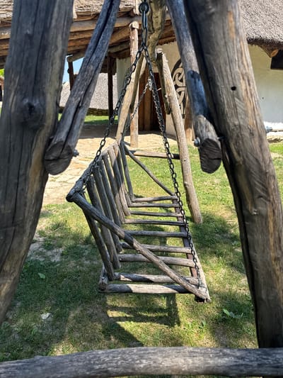The image depicts a rustic wooden swing, constructed from weathered logs and chains, set against a backdrop of traditional thatched-roof buildings. The swing's seat is formed by a series of wooden slats supported by a rough wooden frame. Two large, vertical logs, part of the swing's support structure, frame the foreground, with a horizontal log visible at the very bottom of the frame. The ground around the swing is covered in sparse grass and some patches of dirt. In the background, a building with white walls and a thatched roof is visible, along with another thatched structure to its left. A circular, black and white decorative motif is visible on the white wall of the building. The overall impression is one of a historical or ethnographic setting. This scene is located in the ethno-village "Happy Kingdom" in the city of Indjija Municipality, Serbia. There are no people visible in the image.