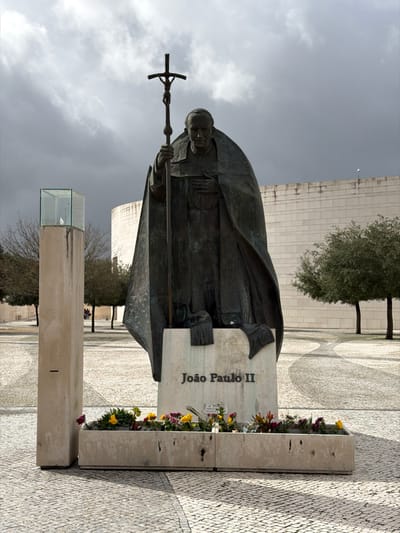 A large bronze statue of Pope John Paul II (João Paulo II) stands prominently in an outdoor paved plaza within the city of Fátima, Portugal, likely at the Sanctuary of Fátima. The statue depicts the Pope cloaked in vestments, holding a tall processional cross with a crucified figure in his right hand, while his left hand rests on his chest. The statue is mounted on a light-colored stone pedestal inscribed with "João Paulo II." In front of the pedestal, two connected rectangular stone planters hold an assortment of colorful flowers (yellow, purple, red) and green foliage, with small notes or offerings placed among them.
To the left of the statue, a tall, slender light-colored stone pillar is visible, topped with a transparent glass enclosure containing a white, possibly avian, object. Further left, several bare-branched trees are sparsely distributed, with two indistinct human figures visible in the distant background. To the right and behind the statue, a substantial, modern building with light-colored, horizontally segmented stone walls and a curved section occupies the background. Several trees with green foliage are positioned between the statue and the building. The entire visible ground is paved with light-colored cobblestones forming subtle patterns. The sky overhead is heavily overcast with dark clouds.