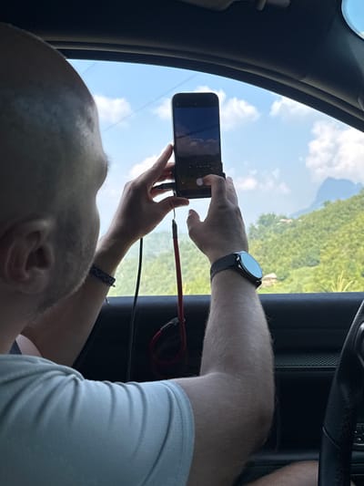 The image shows a person inside a vehicle, possibly a car, taking a photo with their smartphone. The person is male, with a shaved head and fair skin. He is wearing a light-colored shirt and a wristwatch on his left wrist, and a bracelet. The smartphone is black and displays an image. A red and black cable is plugged into the phone. Through the vehicle window, a landscape can be seen, potentially the mountainous terrain found in Mậu Duệ Commune, Vietnam. The presence of a steering wheel indicates the interior is of a car.