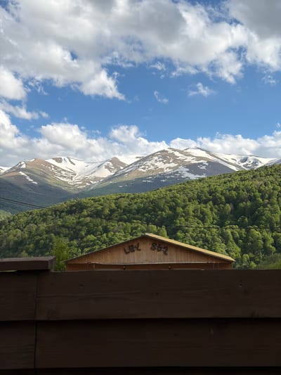 The image depicts a scenic landscape in Lermontovo, Armenia. In the background, there are snow-capped mountains and a dense green forest. A wooden structure, possibly a building, is visible in the foreground. The sky is blue with scattered white clouds. There are no people visible in the frame. Power lines are strung across the landscape. The image is rotated 90 degrees clockwise.