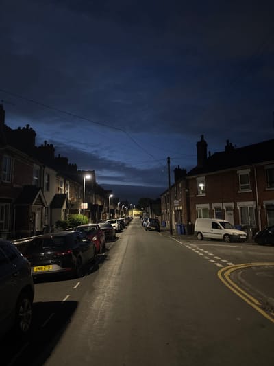 The image depicts a residential street in Stoke-on-Trent, United Kingdom, at dusk. The scene is dominated by terraced houses lining both sides of the street, with parked cars filling the available spaces along the road. Streetlights cast a warm glow on the asphalt, illuminating the parked vehicles and the pavement. The sky above is a deep blue, with scattered clouds suggesting the transition from day to night. The overall atmosphere is quiet and still, characteristic of a residential neighborhood in the evening. There are no people visible in the image. The houses appear to be made of brick, with typical pitched roofs and chimneys. Some windows are lit, suggesting that residents are at home. The street is paved and appears to be in good condition. The parked cars are a variety of makes and models, indicating a typical urban setting.