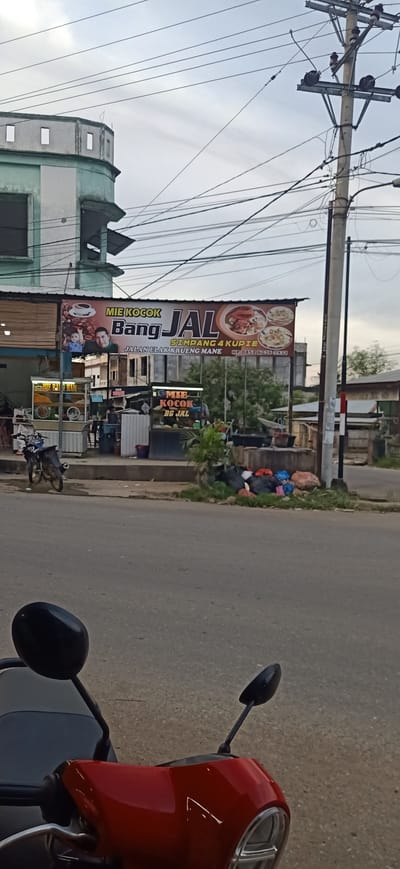 The image shows a street scene in Gampongteungoh, Indonesia, with a red scooter in the foreground and a food stall named "MIE KOCONK BANG JAL" in the background. The food stall has a large banner displaying images of food and text in Indonesian. There are several people visible at and around the food stall, including a child and two adults in the banner. Behind the stall, there are more buildings and trees. A utility pole with numerous wires is also prominent in the scene. The street is paved, and there is a patch of grass with trash bags next to the stall. The sky is overcast.