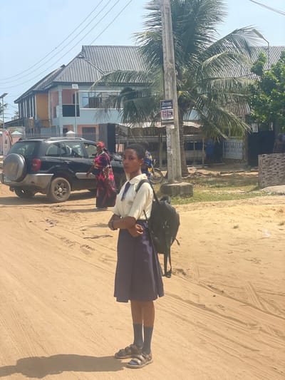 A young girl in a school uniform and sandals is standing on a sandy road in Ikoga, Nigeria. She has a backpack on and is looking at the camera. Beside her is a dark-colored SUV with an older woman seated inside. A palm tree and buildings are in the background. The location appears to be residential. The road is unpaved.