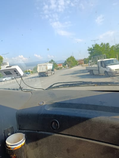 The image shows a view from inside a vehicle, looking out through the windshield. The dashboard is visible in the foreground, with a toll transponder device mounted on the windshield. Outside the vehicle, a road is visible, with several vehicles, including a dump truck and a flatbed truck. Trees and buildings line the road, and mountains are visible in the distance. The sky is blue with some clouds. Based on the presence of the "TollPass" device, it is likely that this image was taken on a toll road. The location is identified as Strumyani, Bulgaria.
