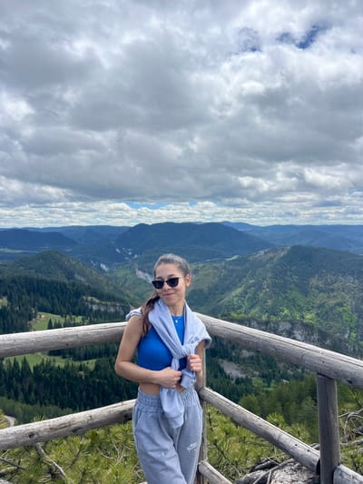 A woman is standing on a wooden observation deck overlooking a mountainous landscape in Giovren, Bulgaria. The woman has sunglasses, a blue top, and a gray tracksuit. She is holding a gray jacket or sweatshirt in her hands. The background consists of layered mountain ranges covered in green vegetation, extending into the distance. The sky above is filled with a mix of white and gray clouds. The wooden deck is constructed with round wooden posts and railings. Some evergreen trees are visible near the railing of the deck.