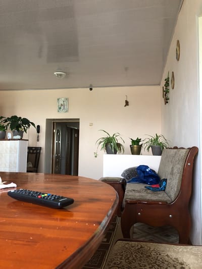 The image shows an interior scene, likely a living room or dining area, with a warm, neutral color palette. In the foreground, a polished wooden dining table occupies the lower left quadrant, with a black remote control with colored buttons resting on its surface. To the right, a wooden bench or sofa with upholstered cushioning and decorative wooden trim is visible. Several potted plants are placed on a white shelf or ledge against the wall. A doorway leading to another part of the house is visible in the background. Decorative elements, including framed pictures and a clock on the wall, add to the domestic setting. The ceiling is a light color, with a visible light fixture. The overall impression is one of a lived-in, comfortable home. There are no people or animals visible in the image.