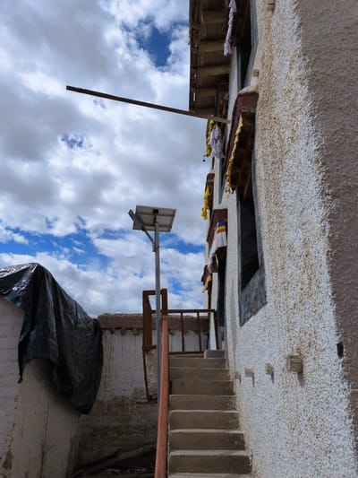 The image depicts an exterior view of a weathered, multi-story building, likely located in Choma, India. The building features a textured, light-colored facade with prominent staining and discoloration. A set of concrete stairs ascends along the side of the building, leading to a small landing area with a rusty metal railing. Above the stairs, a solar-powered street lamp stands on a metal pole. Prayer flags, predominantly white and yellow with some red and blue accents, are visible, hung from the building's upper levels and beneath an overhang. The roof structure appears to be made of wooden beams and corrugated material. In the foreground to the left, a large object covered with a black tarp is partially visible. The sky above is a mix of bright blue with scattered white and grey clouds. The overall scene suggests an outdoor environment, possibly a courtyard or alleyway, adjacent to a traditional or older structure.