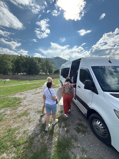 In Kupchegen, Russia, a white passenger van is parked on a gravel and grass lot. Three women are near the open side door of the van. One woman, wearing a white t-shirt and blue shorts, is facing away from the camera and looking towards the van. Another woman, in a floral shirt and orange pants, is also facing the van and appears to be about to enter. A third woman, in a white top and light-colored shorts, stands near the van, facing away from the camera. The van has tinted windows and a side step. In the background, a wooden fence encloses a grassy area with scattered posts, and beyond that, a dense line of green trees and a distant mountain range under a blue sky with scattered white clouds.