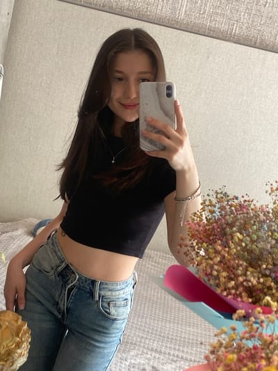 A young woman with long, dark brown hair, wearing a black crop top and light-wash jeans, takes a selfie in a bedroom. She is holding a white and grey marbled phone in her right hand. A small, silver necklace with a star pendant is visible on her neck. She is also wearing a silver bracelet on her left wrist. To her right, a bouquet of small, colorful dried flowers is arranged in a blue and pink wrapping. The background consists of a textured, light-colored wall. The woman is seated on a white bed with a textured surface. The image appears to be taken indoors. The location is Nizhnyaya Maktama, Russia.