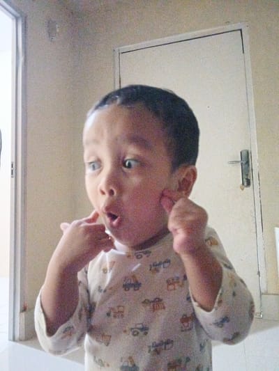 A young child, likely a toddler, stands indoors in front of a white door with a silver handle. The child has dark hair and is wearing a light-colored shirt with a pattern of colorful cartoon vehicles. Their eyes are wide and looking to the left, and they are touching their cheeks with both index fingers, giving them a surprised or playful expression. The background is a plain wall, with a doorway visible on the left side of the frame. The lighting appears to be natural, coming from the left. The overall scene suggests a casual, domestic setting in Cibungbulang, Indonesia.