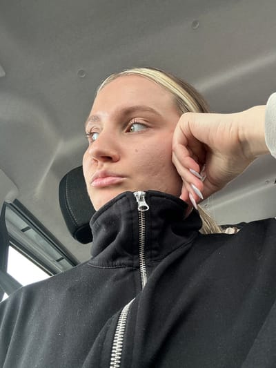 A young adult female is depicted in a medium close-up, situated within the interior of a vehicle in Kırkağaç, Turkey. She is positioned facing left in profile, with her gaze directed outwards through a window that shows a blurred, light-colored exterior. Her hair is blond with lighter streaks, and her eyes are light brown/hazel. A small silver ring is present in her right nostril. She wears a black, high-collared sweatshirt or jacket with a prominent silver zipper, partially open at the neck. Her left hand, featuring long, pointed, light-colored artificial nails, rests against the left side of her face, near her jawline. A black vehicle headrest is visible behind her, and the light gray ceiling of the vehicle fills the upper background. No other individuals or specific landmarks of Kırkağaç are discernible within the frame.