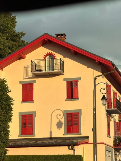 The image shows the exterior of a light yellow building with a red roof and red shutters on its windows. The building has a balcony with a black railing on the second floor, and a similar balcony on the right side. A lamppost with an ornate curved arm and a lantern stands in front of the building. Shadows of the lamppost and tree leaves are cast on the building's facade. The sky is overcast and appears grey. The building appears to be in a town or city setting, possibly in Saint-Gervais-les-Bains, France, given the architectural style and the presence of a lamppost. The overall scene is that of an urban building facade, captured during daylight under a cloudy sky.