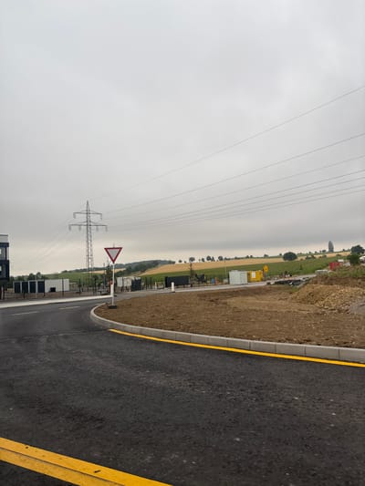 This is an outdoor, eye-level, full-shot photograph of a construction site with agricultural fields and power lines in the background. The sky is overcast and gray. In the foreground, a curved asphalt road with yellow lane markings is visible. To the right of the road, a curb separates the road from a patch of dirt and gravel, with a pile of construction debris. Further back, a power line tower stands tall, with multiple power lines stretching across the frame. Several temporary buildings, likely at a construction site, are also present. The background features rolling green and yellow fields, suggesting agricultural land. The scene is likely located near Untermünkheim, Germany, given the context. There are no people visible in the image.