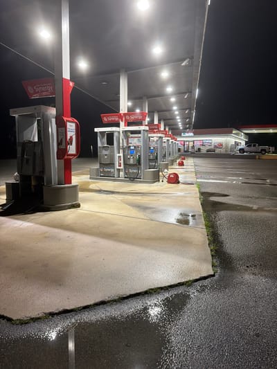 The image shows a gas station at night in Wildwood, United States. The gas station features multiple pumps under a lit canopy. Several red hoses with reels are visible on the ground near the pumps. The ground is wet, with puddles reflecting the overhead lights. In the background, a convenience store is visible with lights on inside and a white pickup truck parked in front of it. The scene appears to be deserted.