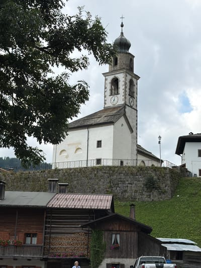 The image shows a view of the town of Sauris di Sopra / Plozn / Sauris Disore, Italy, featuring a white church with a prominent bell tower. The church is situated on a stone-walled embankment, with a dark green tree partially obscuring the left side of the frame. Below the embankment, traditional wooden buildings with visible firewood stacks are present. A white pickup truck is parked in front of one of the buildings, and a person wearing a blue shirt is standing near it. The sky is overcast with scattered clouds.