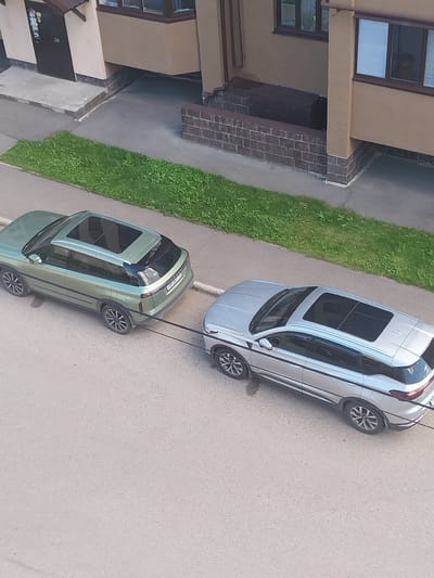 In the city of Salavat, Russia, two SUVs are parked on an asphalt road adjacent to a building with a light brown facade and a brown roof. A thin strip of green grass runs along the side of the road, separating it from the building's foundation, which is decorated with dark brickwork. The first SUV, a light green vehicle, is parked on the left, facing away from the camera. A black towing cable extends from its rear bumper to the front bumper of the second SUV, a silver vehicle, which is parked on the right, also facing away from the camera. Both vehicles have sunroofs.