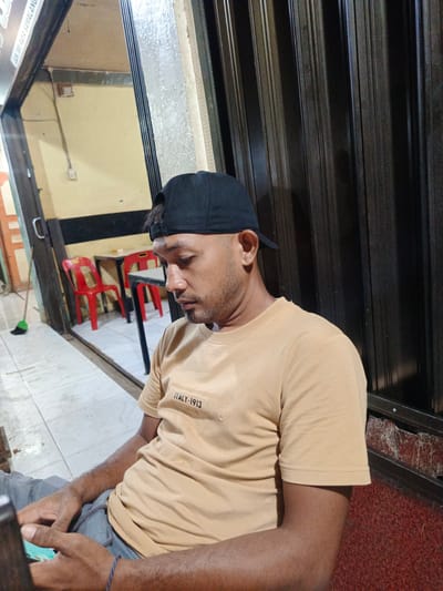 An adult male, wearing a black baseball cap, a light brown short-sleeved t-shirt with "ITALY-1913" text, and grey drawstring pants, is seated and looking downwards at a smartphone held in his hands. A blue beaded bracelet is visible on his left wrist. The scene takes place in Sangso, Indonesia. The background to the man's right features a dark, vertically corrugated metal surface. To his left, through an open doorway, an interior space with light yellow walls is visible, furnished with two red plastic chairs and two tables with dark tops. The floor consists of light grey square tiles. A brown broom with a green base leans against the wall. Partial signage above the doorway reads "JLN SRI RAYA SAMALANG".