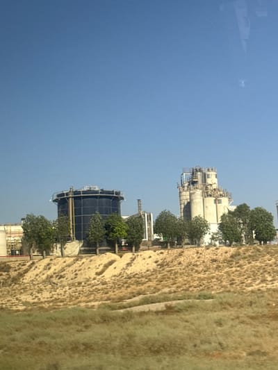 The image depicts an industrial scene in Arad, Israel, under a clear, bright sky suggesting daytime, possibly morning or afternoon. The main subjects are industrial structures, including several large, cylindrical storage tanks and a complex of interconnected towers and pipelines. One prominent tank is dark blue and appears to be covered with a mesh netting. Another, taller structure features multiple white, cylindrical tanks of varying sizes, connected by a network of scaffolding and pipes. These structures suggest an industrial or processing facility, likely related to petrochemicals, water treatment, or a similar industry.
The setting is a dry, somewhat arid environment, as indicated by the yellowish, rocky terrain bordering the industrial site. Patches of green, scrubby vegetation and trees are interspersed throughout the scene, providing a contrast to the man-made structures and the dry earth. The overall impression is one of industrial activity within a natural landscape.
There is no visible text in the image. The image captures a static view of the industrial complex and its surroundings, with no apparent human activity or specific event taking place. The lighting suggests a sunny day, and the absence of clouds indicates fair weather. The perspective is from a slightly elevated viewpoint, looking across the landscape towards the industrial facilities.
