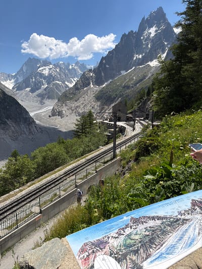 This image depicts a scenic mountain landscape in Chamonix-Mont-Blanc, France, featuring a cogwheel railway track winding through the terrain. In the foreground, a detailed map of glaciers and mountain summits is laid out. A person is visible walking along a path next to the railway tracks, equipped with a backpack and walking poles. The tracks themselves are bordered by a fence and a concrete wall. Beyond the tracks, lush green vegetation covers the mountainside, with a vast glacial valley and snow-capped peaks dominating the background under a clear blue sky with scattered clouds. A stone building, possibly a station, is situated alongside the railway line further up the track.