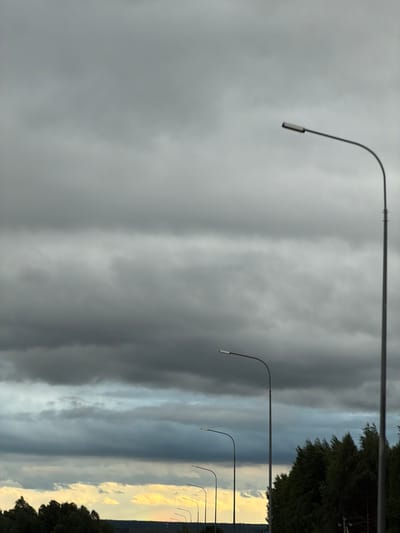 The image shows a row of streetlights lining a street in Крынды, Russia. The sky is predominantly gray and cloudy, with varying shades of gray and some lighter areas near the horizon. A line of trees or bushes is visible on the right side of the frame. The streetlights are simple in design, with curved arms extending from the poles. The image has been rotated.
