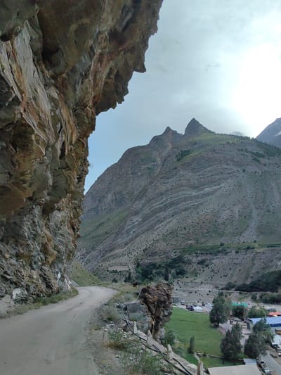 The image depicts a dramatic mountainous landscape with a dirt road winding through a valley. On the left, a steep, rocky cliff face overhangs the road, creating a sense of enclosure. The road is empty of vehicles. Beyond the road, the valley floor opens up to reveal a small settlement with buildings and green fields. In the distance, imposing mountains rise, their slopes covered in a mix of rock, sparse vegetation, and some scattered trees. The sky above is overcast, with diffused sunlight. The location is identified as Gushal, India.
