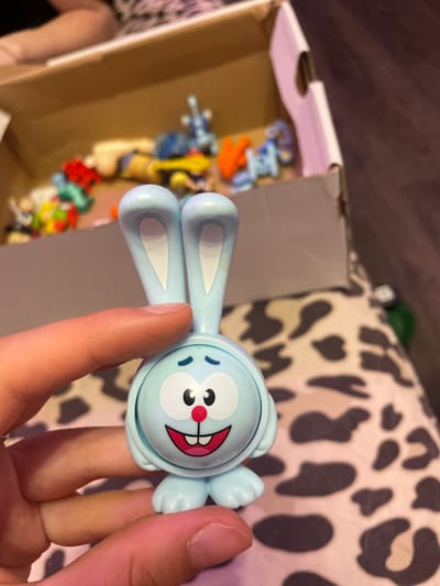 A close-up shot shows a light blue, cartoonish bunny figurine with large ears, wide eyes, and an open mouth with visible teeth and a red tongue, being held by a person's hand. The figurine is the primary subject in the foreground. In the background, a cardboard box is visible, filled with other colorful, small toys. The surface beneath the box appears to be a patterned fabric, possibly a blanket or upholstery, with a leopard-like print. The lighting suggests an indoor setting. There is no discernible information in the image to link it to Borisova Griva Massiv, Russia.