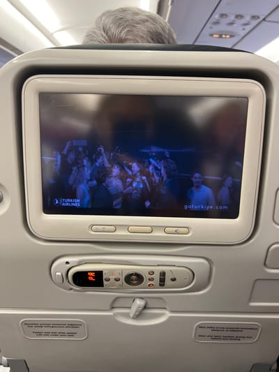 The image displays the interior of an aircraft cabin, specifically the view from a passenger seat, within an aircraft located in the city of Steenokkerzeel, Belgium. The central focus is the in-flight entertainment (IFE) screen mounted on the seat back in front of the viewer.

The IFE screen, framed in white plastic, exhibits minor surface scratches and reflections. It displays a video showing a group of approximately fifteen individuals engaged in activities resembling dancing or socializing in a dimly lit, dark blue-hued environment, possibly a concert or club setting. Visible on the screen are the "TURKISH AIRLINES" logo in the lower-left corner and the URL "goturkiye.com" in the lower-right.

Beneath the screen, an integrated control unit for the IFE system is present. It features a grey remote control with multiple buttons for various functions (e.g., volume, channel navigation, power) and a small orange-illuminated digital display showing the characters "HI". A white plastic latch for a tray table is positioned directly below this control unit.

Two rectangular informational stickers are affixed to the seat back below the tray table latch. The left sticker provides instructions in Turkish and English regarding fastening seatbelts and locating life vests ("Fasten seat belt while seated / Life vest under your seat"). The right sticker instructs passengers in Turkish and English to stow and latch the handset during taxi, takeoff, and landing ("Stow and latch handset during taxi, takeoff and landing").

The top portion of the image shows the back of an individual's head, characterized by short, neatly combed grey hair, indicating an adult passenger in the seat directly ahead. Above this, the aircraft's white overhead panel and cabin lighting fixtures are visible, with a small orange indicator light illuminated. The cabin interior is predominantly white and light grey.