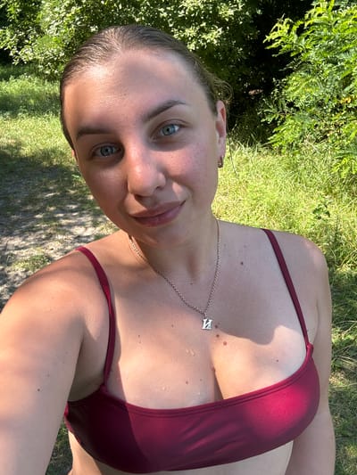 The image is a close-up, waist-up selfie taken outdoors during the day. A woman with fair skin and light blue eyes is positioned in the foreground, looking directly at the camera. She has her hair pulled back and is wearing a maroon bikini top with thin straps. A silver necklace with a letter pendant rests on her décolletage. Water droplets are visible on her skin and bikini top. The background consists of green foliage and grass, with dappled sunlight and shadows suggesting a wooded or park-like area. The location is identified as Chykalivka, Ukraine.