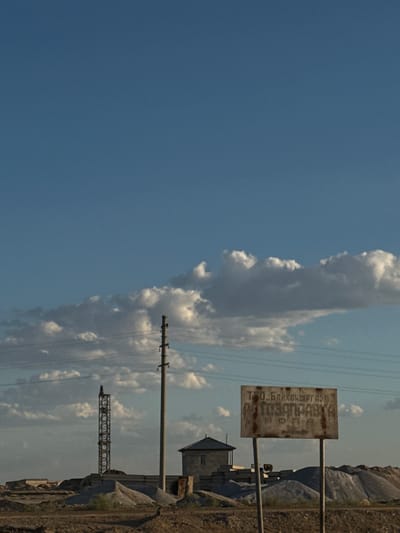 The image depicts a desolate landscape under a clear blue sky with scattered clouds. In the foreground, a rusted metal sign with Cyrillic text is mounted on a pole. The text on the sign appears to read: "ТОО. БАЖХОВГАЗНЕФТЕПРОДУКТ" and below it, "ТОО. ТУГАНБАЙ". This suggests a business or company name related to gas and oil products.

To the right of the sign, a small, weathered building with a dark roof stands against a backdrop of rugged, rocky terrain. Several power lines stretch across the scene, supported by utility poles. One such pole with multiple insulators is prominently featured. Further in the distance, a large, lattice-like metal structure, possibly part of a power transmission tower, is visible. The overall scene conveys a sense of isolation and industrial presence in a remote, arid environment. The lighting suggests it is daytime, likely late morning or early afternoon, with the sun casting subtle shadows. The weather appears fair, with a gentle breeze indicated by the clouds. The location is Töretam, Kazakhstan, known for its harsh climate and sparse vegetation. There are no people visible in the image.