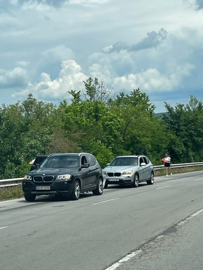 The image shows two BMW X1 cars parked on the shoulder of a road. The first car is black with the license plate "AG-01-AYN." The second car is silver with the license plate "AG-21-EWS." A person wearing a white shirt and dark shorts stands next to the silver car, facing away from the camera. The background includes green trees and a cloudy sky. The location is likely near Osenets, Bulgaria based on the license plate prefix "AG" which corresponds to the region.