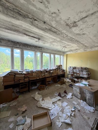 The image depicts an abandoned room, likely a former office or archive, within a building in Nikolsky, Russia. The room is filled with stacks of papers and documents, piled high on desks and on the floor. The papers appear aged and disheveled, with many scattered around the room. The overall environment suggests neglect and disuse, with peeling paint on the ceiling and walls, and debris on the floor. Through the large windows, a dense forest can be seen, indicating the building's location outside of a dense urban area. There are no people visible in the scene.