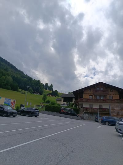 The image depicts an overcast day with a cloudy sky over a mountainous region. In the foreground, a paved parking lot with white lane markings is visible, with several cars parked. To the left, a dark-colored SUV and a dark-colored sedan are parked. A black van with text and logos is parked on an incline to the left of the building. In the middle ground, a wooden building with multiple balconies and windows is situated on a grassy slope. Further to the left, a lush green hillside covered with trees rises steeply. A ski lift with chairs suspended from cables ascends the slope. In the distance, more buildings and trees dot the landscape. The scene is likely a parking area at the base of a ski resort or a similar mountain recreation area, possibly in or near the city of Hallein, Austria, given the presence of the ski lift and alpine architecture.