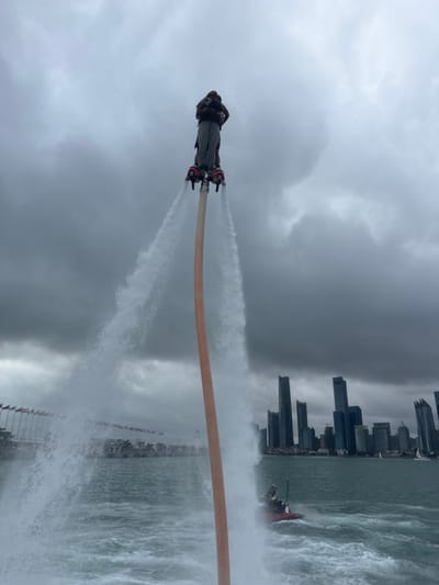 A person is captured airborne on a flyboard device over a body of water in Shinan District, Qingdao, China. The individual, dressed in dark clothing and a helmet, is elevated by two powerful jets of water emanating downwards from foot-mounted boots, which are connected to a thick, light-colored hose extending into the water below. To the right, a red and black jet ski with an operator is actively moving, generating a wake, and is responsible for providing the water pressure to the flyboard system. The background features a dense skyline of modern skyscrapers, dark in hue, rising from the distant shoreline. To the left, a long series of colorful flags is visible along a waterfront structure. The sky is heavily overcast with dark gray clouds. The water surface is choppy. A small white sailboat is discernible in the far distance near the city skyline on the right.