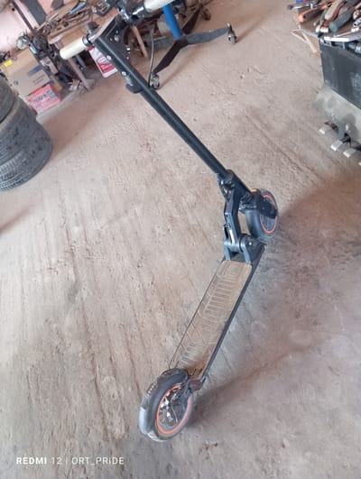 A black electric scooter with orange accents is parked on a dusty concrete floor. The scooter is positioned at an angle, with its handlebars visible in the upper portion of the frame and its front wheel in the lower right. The floor shows various scuff marks and dust accumulation, indicative of an industrial or workshop environment. In the background, to the left, stacks of tires are visible, along with some boxes and tools. To the right, part of a large black object, possibly a piece of machinery or storage unit, is present, with metal pedals attached to its base. The lighting suggests an indoor setting with natural light entering from an unseen source, casting shadows on the floor. The image is watermarked with "REDMI 12 | ORT_PRIDE" in the lower left corner. This scene is likely located in an unpaved workshop or storage area within the city of Kizlyar, Russia, given the presence of tools, tires, and industrial elements, suggesting a location where vehicles or equipment are stored or repaired.