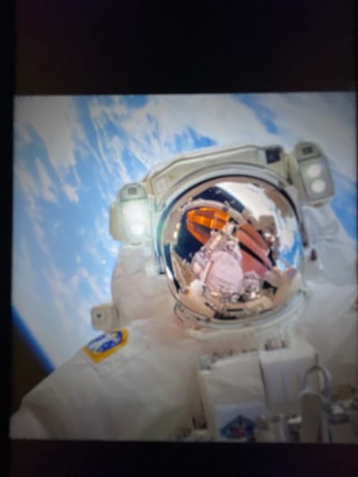 The image shows a close-up view of an astronaut in a spacesuit, with the Earth visible in the background. The astronaut's helmet visor reflects a complex array of equipment and lights, suggesting an interior view of a spacecraft or station. The Earth appears as a blue and white orb with cloud formations. There is no visible indication of Jihlava, Czechia in the image.