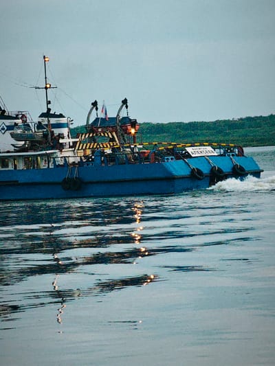 A blue barge with a white and blue superstructure is traveling on a body of water. The barge has a mast with two orange lights, and two flags are visible on the stern. There are yellow and black striped railings along the deck. A white sign on the stern reads "КАПИТАН АЛЕКСЕЕВ" (Captain Alekseev). Three large tires are hanging from the side of the barge, and a smaller boat is attached to the superstructure. The water is rippled, with reflections of lights visible on the surface. The background shows a tree-covered shoreline under a hazy, overcast sky. The scene appears to be set in the early morning or late evening, given the soft light and muted colors. The overall impression is one of a working vessel on a calm waterway, possibly in a rural or industrial setting.