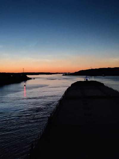 The image captures a twilight scene over a body of water, likely a canal or river, near Sud'bitsy, Russia. A large, dark silhouette of a vessel occupies the foreground, stretching across the right side of the frame. Its deck is flat and indistinct in the low light. The water in the foreground is dark and choppy, reflecting the dim sky.
Further into the scene, the water becomes smoother, reflecting the gradient of the sky. The sky itself transitions from a deep, dark blue at the top to a soft orange and pink hue near the horizon, indicating either dawn or dusk. On the left side of the water, a red navigation buoy with a red light is visible, marking the waterway. In the distance, along the shorelines, silhouetted trees and structures are faintly discernible, including a tall pylon-like structure on the left and what appears to be a lighthouse on a hill on the right. A few small lights from distant buildings or vessels are scattered along the horizon. There are no discernible people or specific actions depicted in the image. The overall mood is serene and quiet, typical of twilight hours.