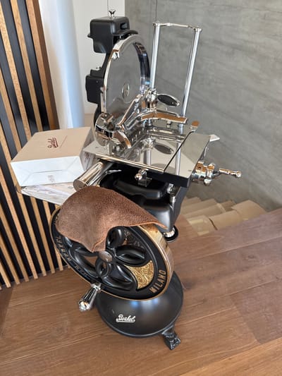 A black and chrome Berkel slicing machine is positioned on a wooden floor. The slicing machine has a large, shiny metal blade, a black base, and a decorative wheel with "BERKEL MILANO" inscribed on it. A brown cloth is draped over the wheel. To the left of the slicing machine, a light brown cardboard box with the "Berkel" logo in red lettering is visible, resting on a clear plastic tray. Behind the slicing machine, a wall with vertical wooden slats in alternating black and brown tones is present. The background also shows a section of a gray concrete wall. The scene appears to be indoors, likely in a home or a commercial space in Masera, Italy.