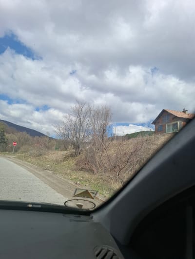 View from inside a vehicle of a rural landscape near Gintsi, Bulgaria. Visible are a road, a stop sign, a bare tree, a hillside with sparse vegetation, a building with a red tile roof, and a cloudy sky. A section of the vehicle's interior is prominent in the right side of the frame.