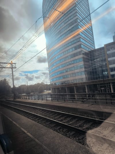 The image depicts a view from a train, looking out at a curved, modern glass-facade office building in Hoofddorp, Netherlands. The train tracks run diagonally across the foreground, with a platform visible to the left. Overhead, electrical wires and support structures for the train line are prominent. The building, which appears to be the "Skytower" or a similar structure at the Siriusdreef area, dominates the right side of the frame. Sunlight streaks across the building's facade, creating bright orange reflections. The sky is overcast with some patches of blue. Trees line the area behind the platform. There are no people clearly visible in the frame. The scene conveys a sense of urban transit and modern architecture.