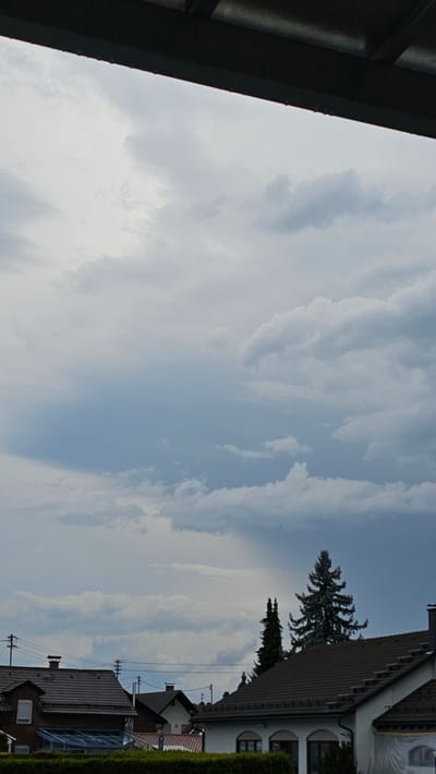 The image depicts a cloudy sky over a residential area in Vöhringen, Germany. A dark overhang is visible at the top of the frame, suggesting a structure like a porch or balcony. Below this, a dense formation of clouds dominates the upper portion of the sky, with lighter clouds scattered throughout. In the lower part of the image, several houses with dark roofs are visible, along with trees, utility poles, and power lines. The lighting suggests overcast conditions, possibly before or during rain.
