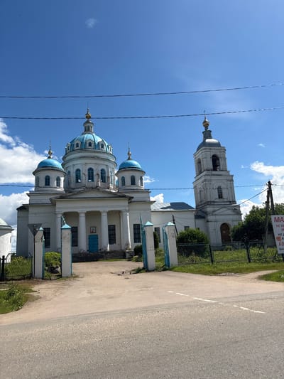 The image shows the Church of the Resurrection of Christ in Novoye, Russia. The church is a white building with a prominent blue-domed central structure and several smaller blue domes. To the right of the main building is a white bell tower with a grey dome. The church is surrounded by a fence and greenery. In the foreground, there is a paved road leading to an entrance gate with blue-topped pillars. The sky is blue with scattered white clouds. A sign is visible on the right side of the image with Russian text. No people are present in the image.
