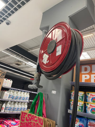 The image shows a fire hose reel mounted on a grey column in a supermarket in Saint-Brice-Courcelles, France (latitude: 49.283, longitude: 4). The reel is red with a black hose wrapped around it. The hose is partially unwound, with the end of the hose lying on a shelf displaying cleaning products. The shelf is orange and white, with a large yellow sign reading "DE PR". The hose is attached to a fire hydrant with a silver valve. There are a number of shopping bags on the shelves below the hose reel. The supermarket ceiling has a white grid pattern and there are overhead fluorescent lights. A security camera is visible in the top left corner of the image. There are no people in the image. The scene appears to be in a supermarket aisle.