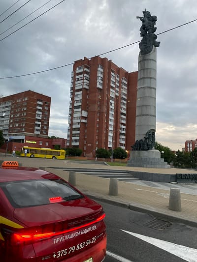 The image depicts a city scene with a prominent monument in the background. The monument is a tall, grey column topped with a sculptural group of figures, possibly representing soldiers or historical figures. To the right of the monument, there is a stone base and steps. In front of the monument are two tall, red brick apartment buildings with many windows. A yellow bus is parked in front of one of the buildings, and a red car, likely a taxi, is in the foreground. The car has text on its rear, including a phone number and what appears to be a slogan. The sky is overcast, suggesting a cloudy day. The overall impression is one of an urban environment, possibly a public square or park area. Visible text on the red car includes "ПРИГЛАШАЕМ ВОДИТЕЛЕЙ" which translates to "WE INVITE DRIVERS," and the phone number "+375 29 883 54 25". There is also text "BEIJING" visible on the car. The location context provided is Лугаваслабадскі сельскі Савет, Belarus.